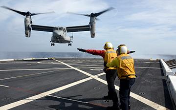 2 men in a flight crew signaling to an MV-22 landing 